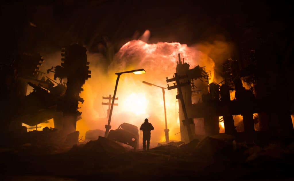 A person walking down a broken road the building around are crumbling and it appears that bombs are exploding in the background. 