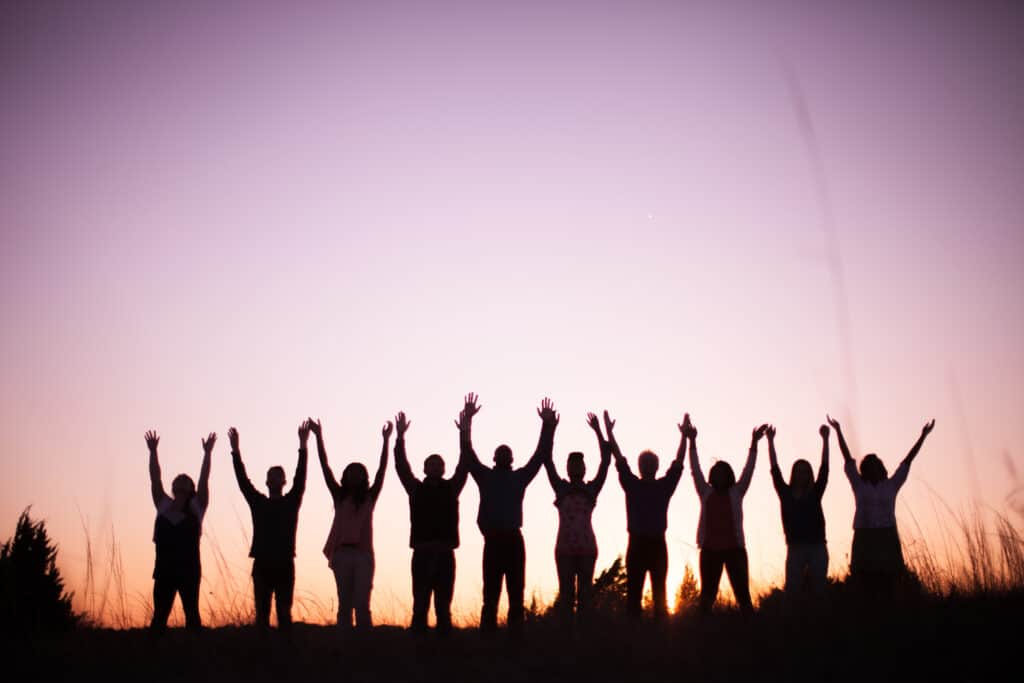 Group of people stand silhouetted with arms raised as the sun sets with a purple sky.