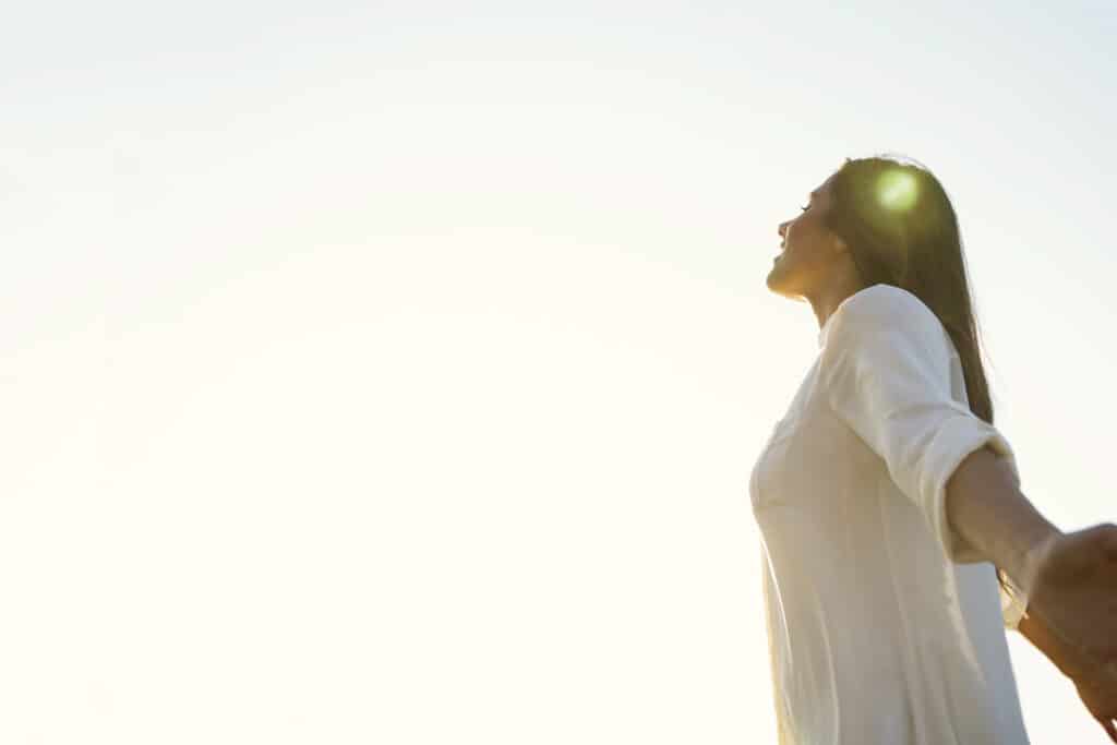 Woman standing with face towards sun and arms outstretched behind her. 
