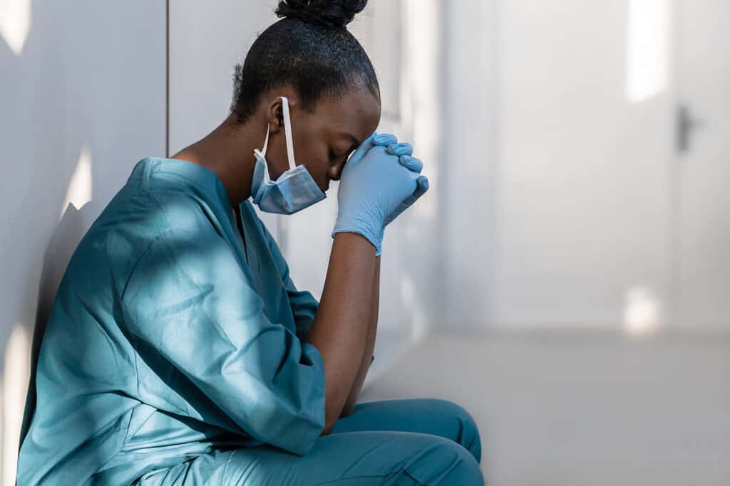 A medical working wearing a mask and gloves sitting on the ground with elbows on knees and head resting on hands. 