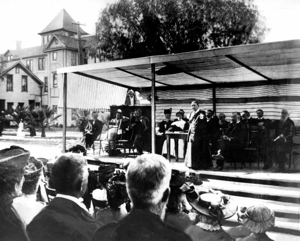 Black and white photo of Ellen White speaking on stage in front of a crowed, with men and women sitting behind her. 