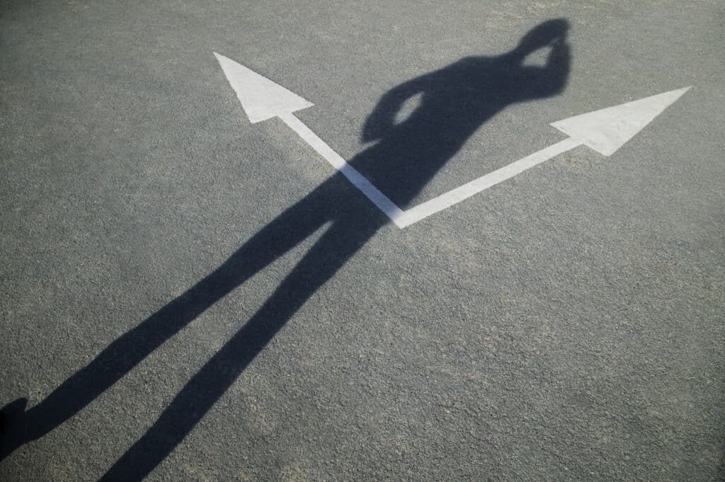 The shadow of a man looking down at a road sign pointing in two different directions, as he makes his choice.