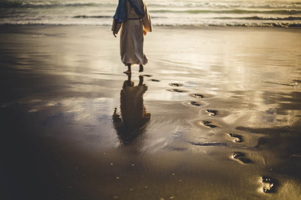 Jesus walking along a beach leaving footprints in the sand, representing how we are to follow His example.