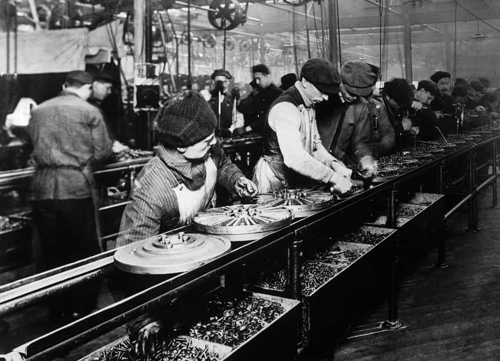 A row of young men work in an unhealthy factory line during the 1900s, assembling mechanical components.