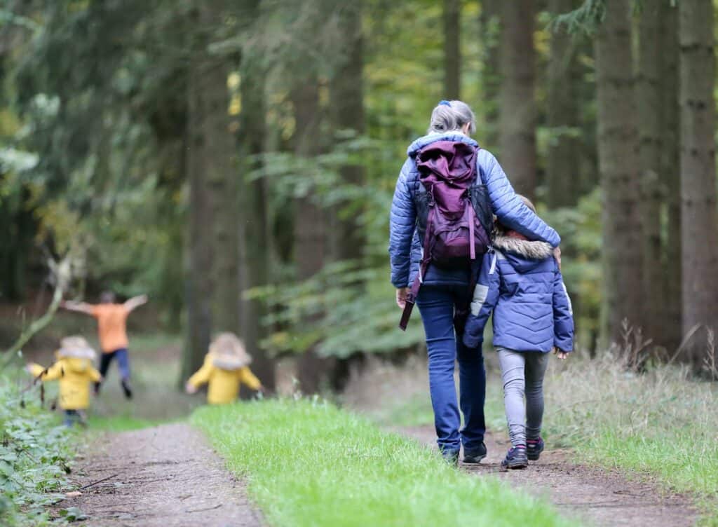 Mom hikes hugging her daughter while walking down a wooded path with her other three children.