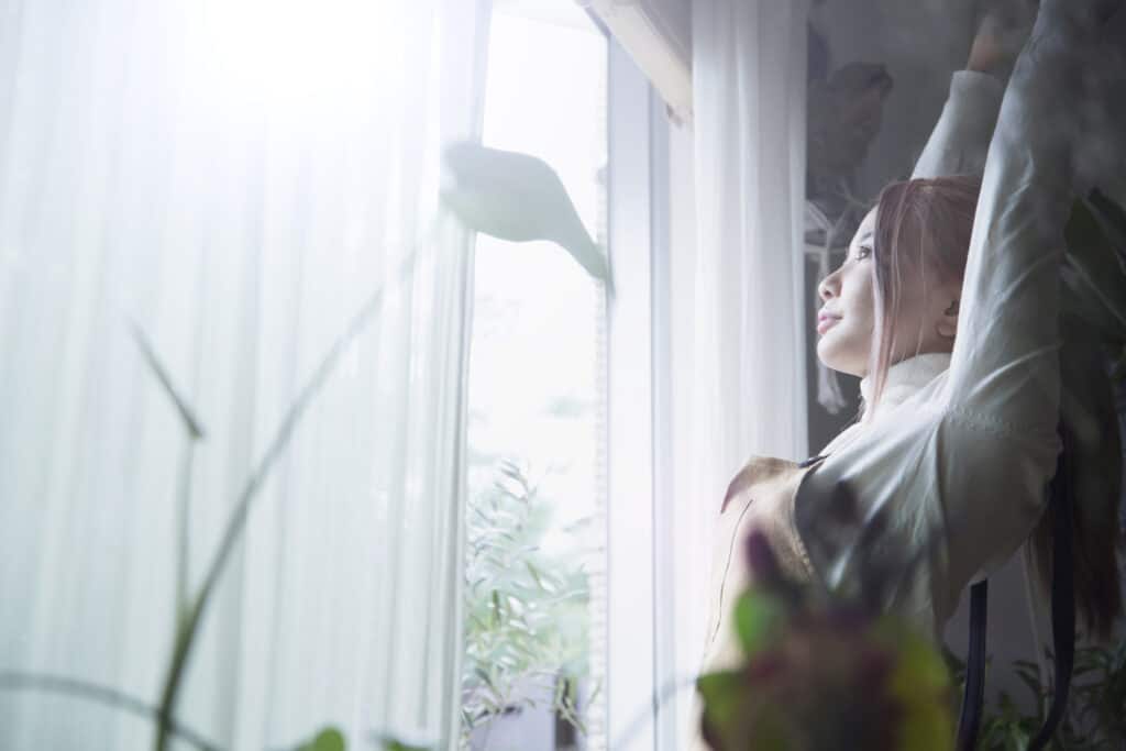 Woman stands in front of a large window soaking in the natural sunlight as she stretches.