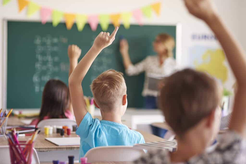 Three students raising their hands as their teacher points to a board math problem.