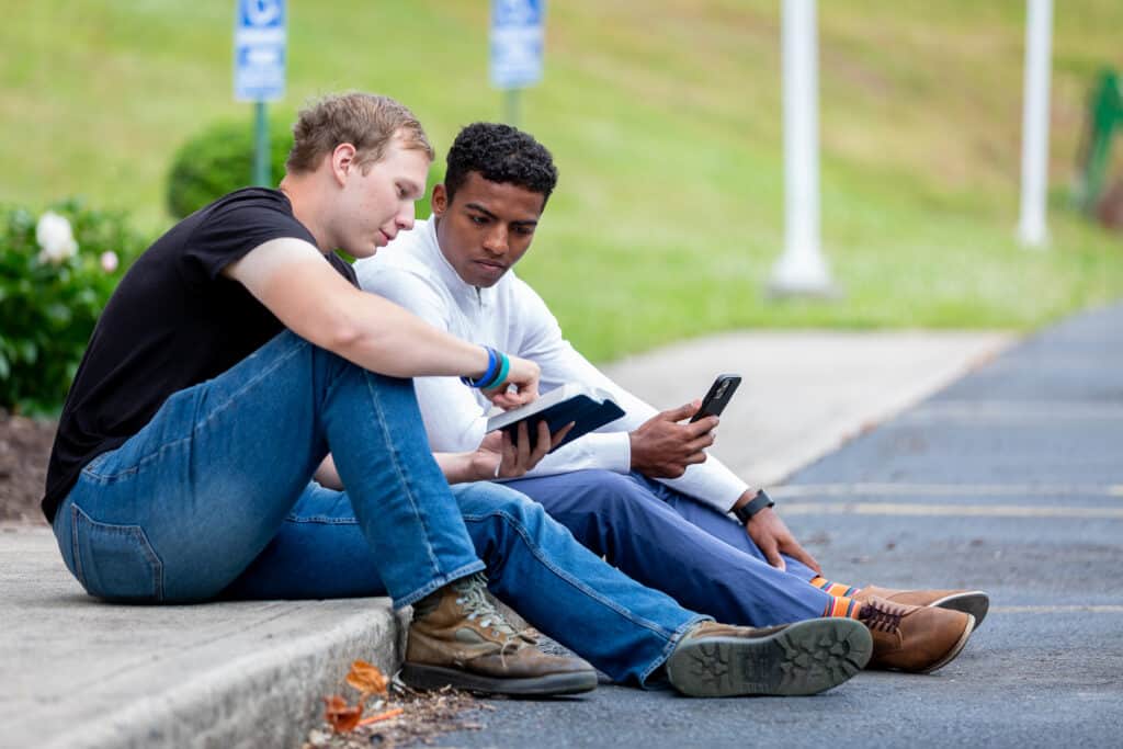 Two men sitting on a sidewalk as one man shows the other a Bible verse.
