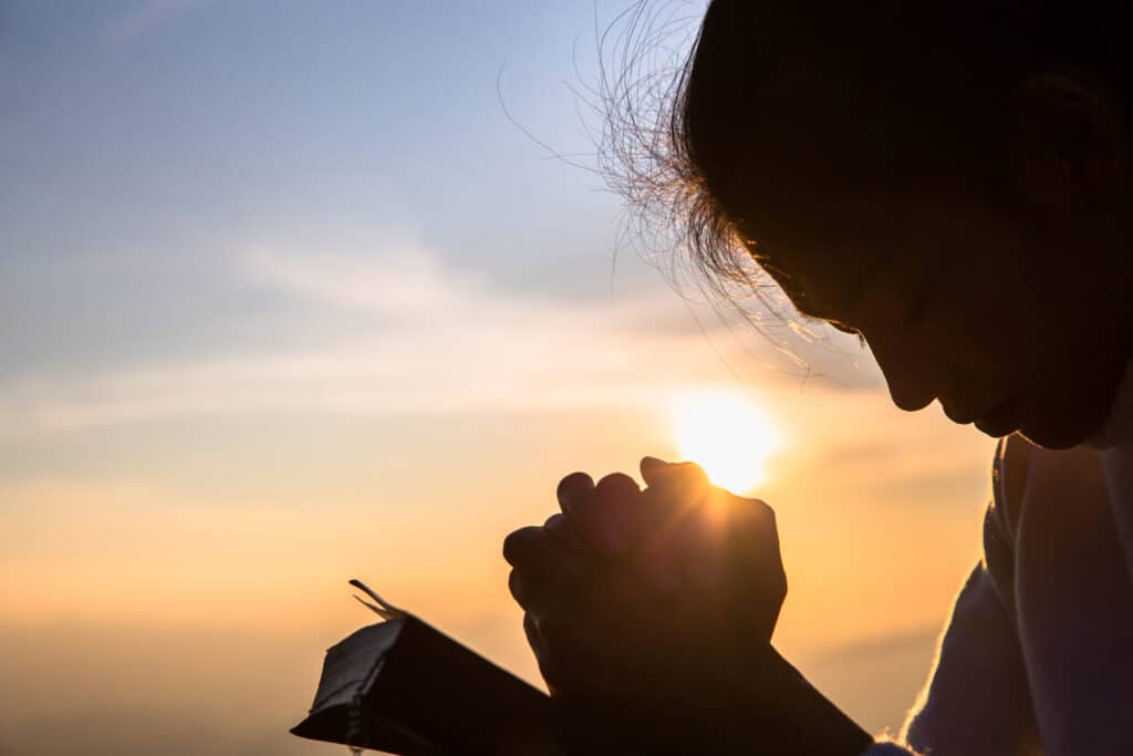 Silhouette of someone praying over a Bible at sunset.