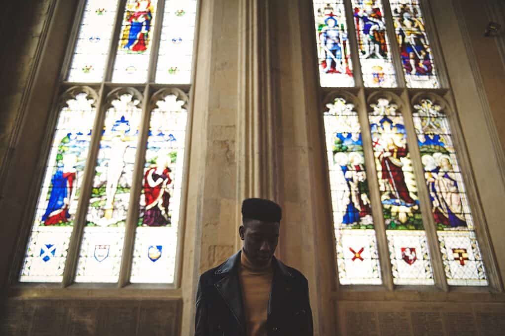 A man sits in front of stained glass windows of a church, taking time to talk with God.