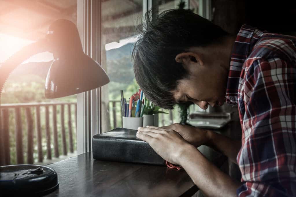 A man is in deep prayer over a Bible, approaching God directly for forgiveness.