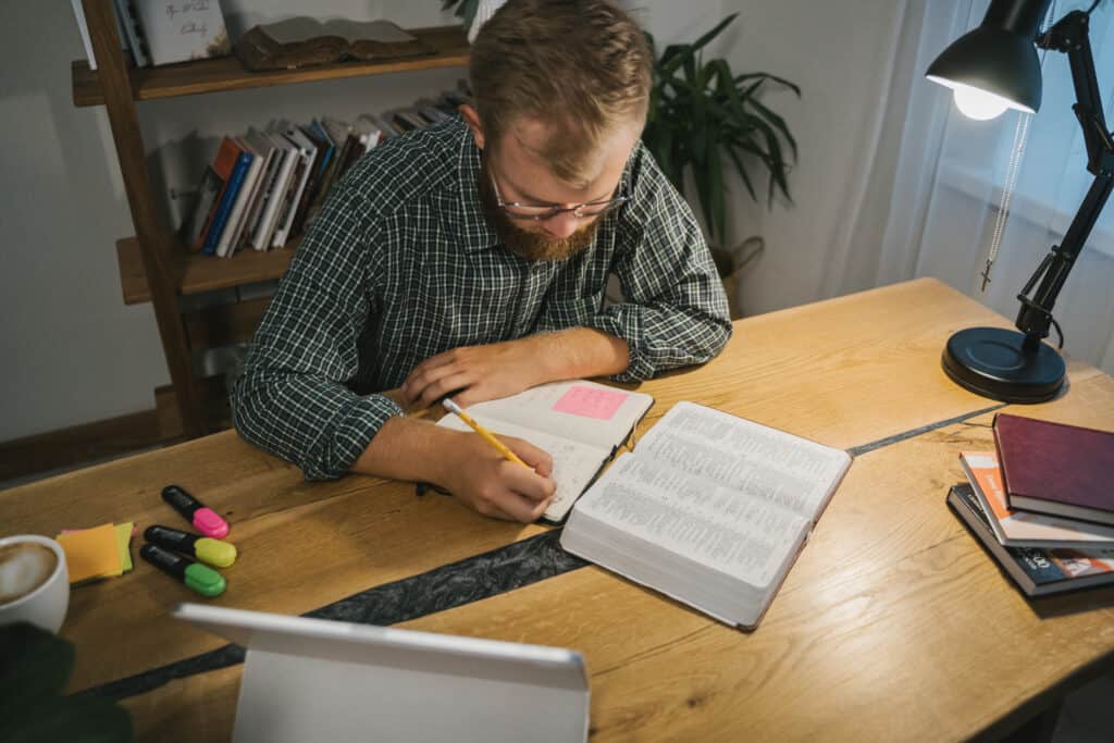 A man takes notes while studying the Bible on a small wooden desk.